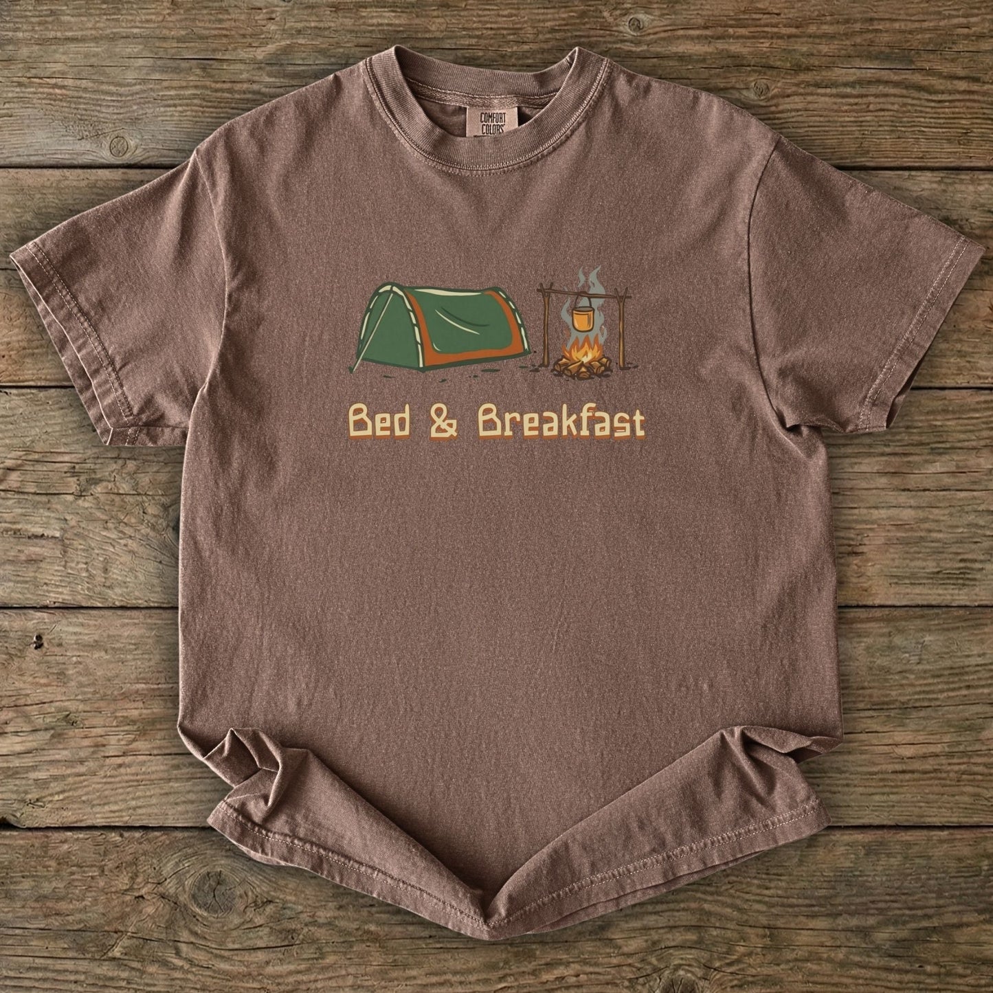 Brown t-shirt with camping graphic and 'Bed & Breakfast' text on a wooden surface