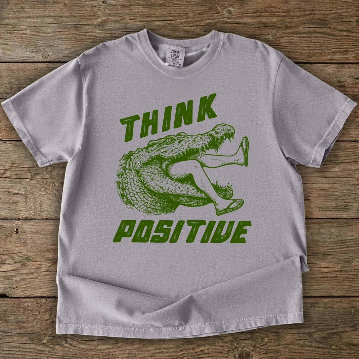 Folded t-shirt with saltwater croc graphic and 'THINK Positive' text on a wooden background