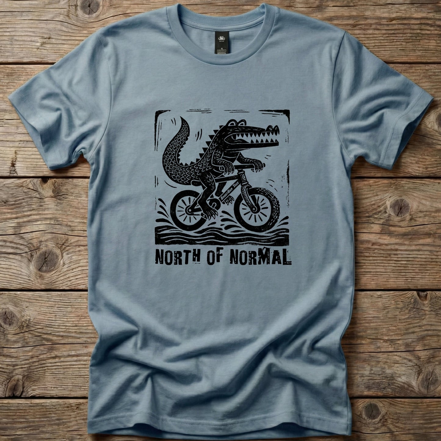 Slate blue Ghost Gum Tees t-shirt featuring a black linocut-style graphic of a crocodile riding a bicycle with the text North of Normal.