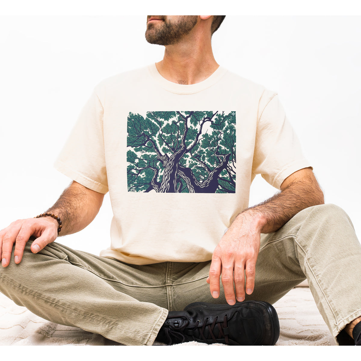 A man wearing a cream-colored t-shirt with a square graphic print of a gnarled tree in dark green and navy blue, paired with khaki pants and black shoes, sitting cross-legged against a white background.