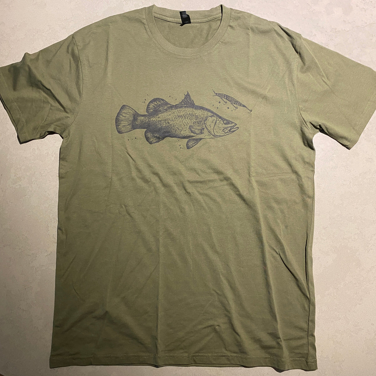 An olive green short-sleeve t-shirt laid flat on a light-colored surface, featuring a dark gray graphic of a barra fish on the chest.