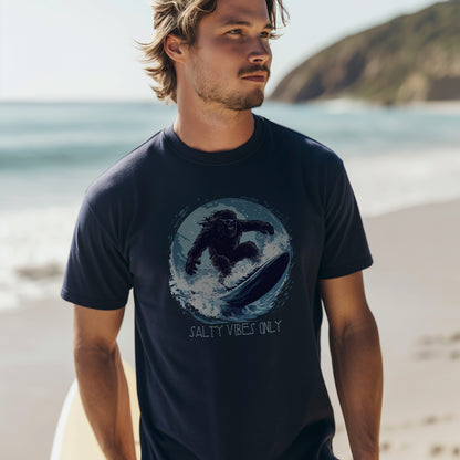 Man wearing navy Bigfoot Surfing T-shirt with "Salty Vibes Only" text, standing on beach with surfboard – lifestyle product mockup