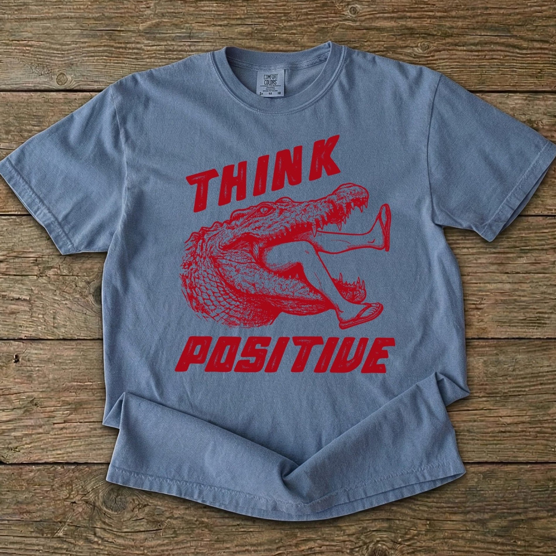 Folded t-shirt with saltwater croc graphic and 'THINK Positive' text on a wooden background