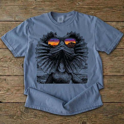 Blue t-shirt with a graphic of a Frilled neck lizard wearing sunglasses on a wooden surface