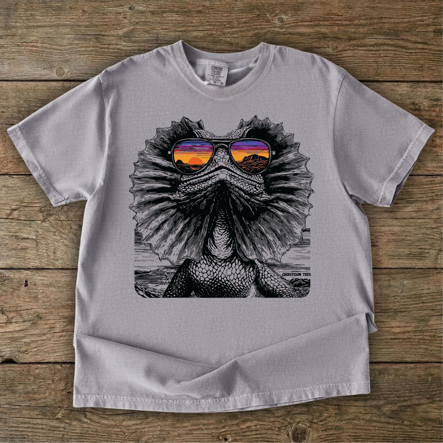 Grey t-shirt with a graphic of a Frilled neck lizard wearing sunglasses on a wooden surface