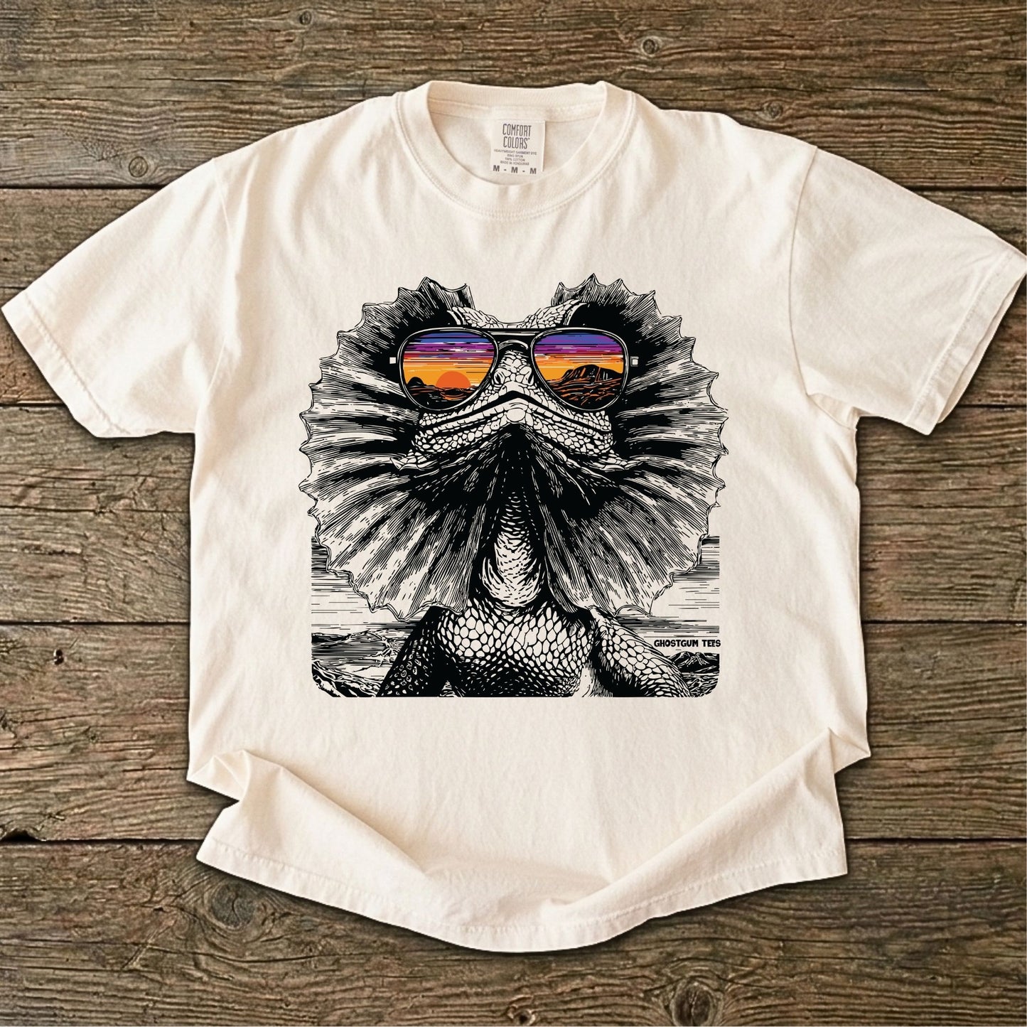 Ivory color t-shirt with a graphic of a Frilled neck lizard wearing sunglasses on a wooden surface