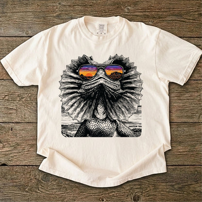 Ivory color t-shirt with a graphic of a Frilled neck lizard wearing sunglasses on a wooden surface