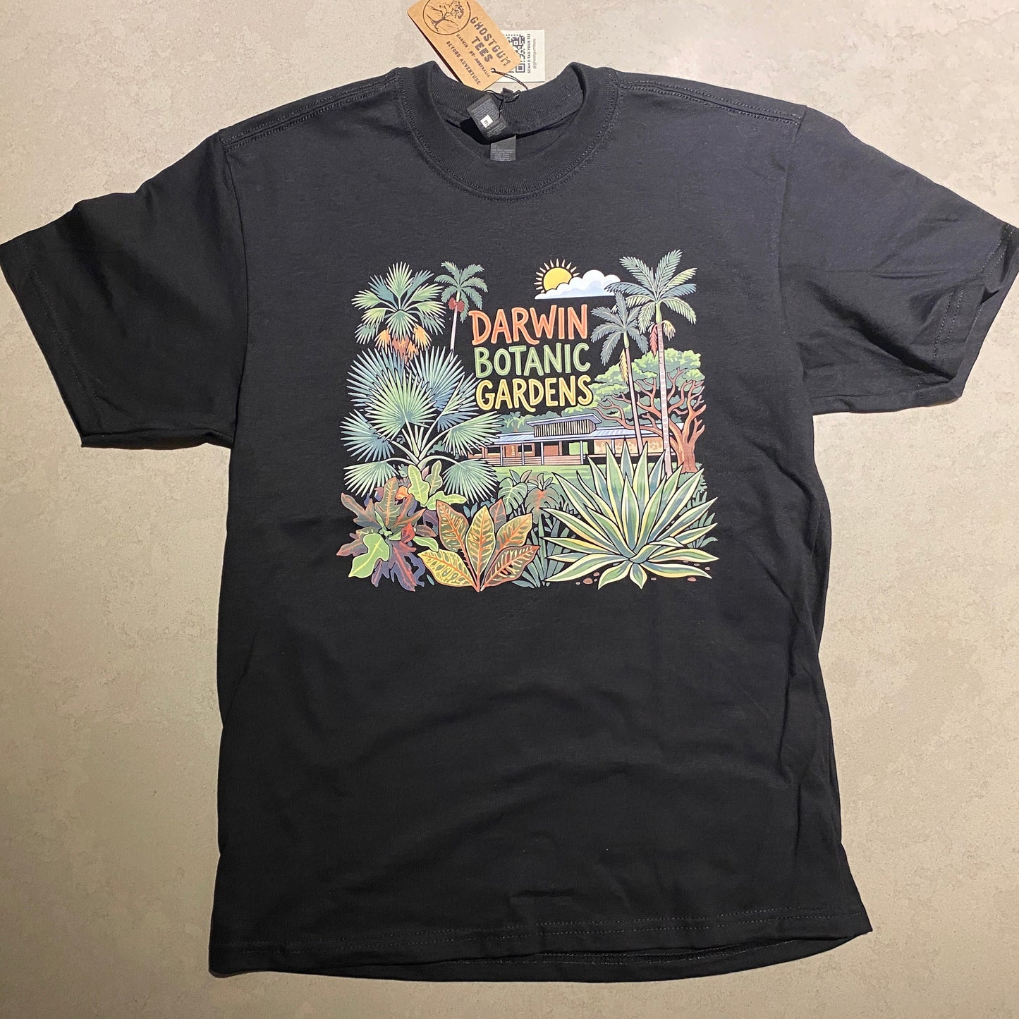 A black short-sleeve graphic T-shirt laid flat on a light beige surface. The front features a colorful tropical botanical design with palm trees, flowers, and vertical text reading 'Tropical Darwin Botanical Gardens.