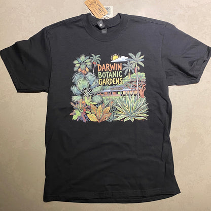 A black short-sleeve graphic T-shirt laid flat on a light beige surface. The front features a colorful tropical botanical design with palm trees, flowers, and vertical text reading 'Tropical Darwin Botanical Gardens.