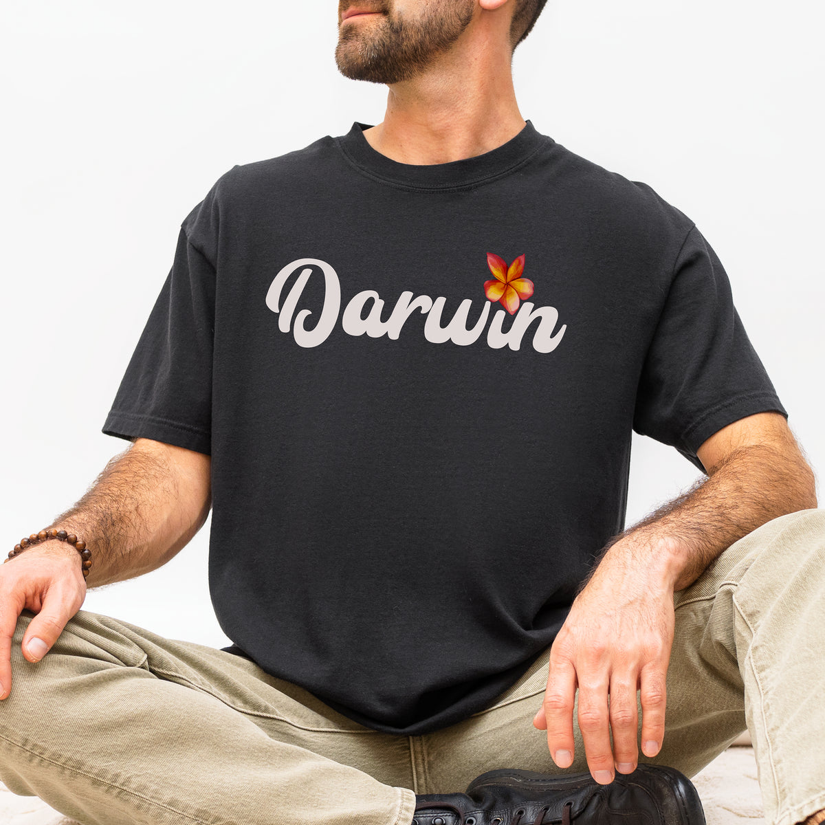 A man wearing a black t-shirt with 'Darwin' written in white script and a small orange and yellow frangipani flower graphic, paired with khaki pants and black shoes, against a plain white background.