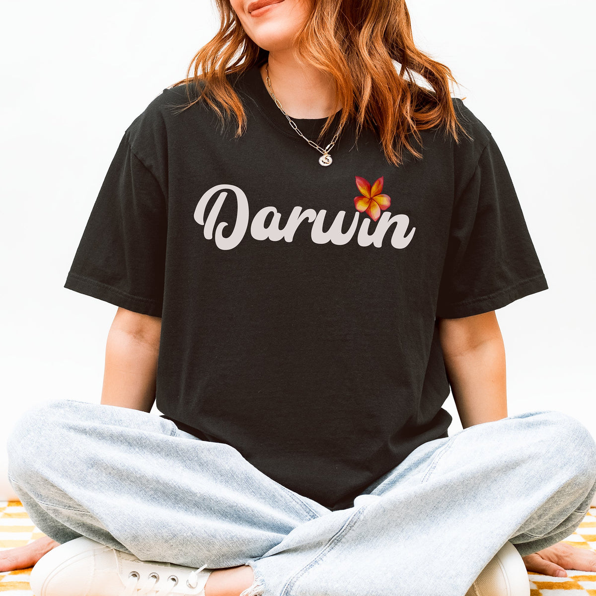 A woman sitting cross-legged wearing a black t-shirt with 'Darwin' written in white script and a small orange frangipani flower graphic, paired with light-wash jeans and white sneakers.