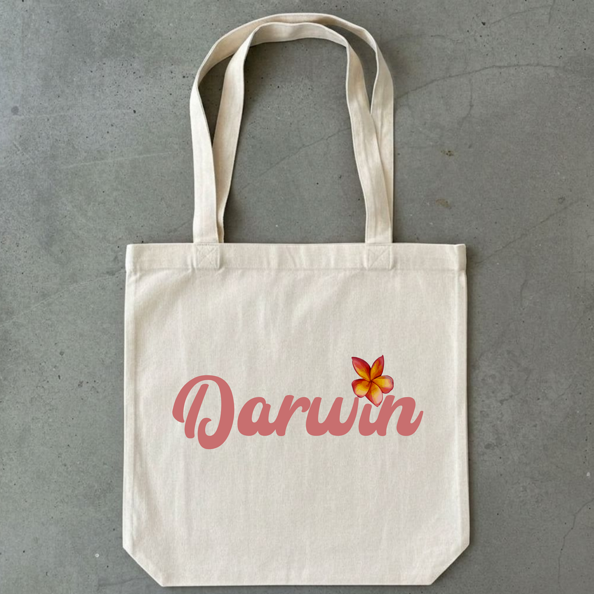 A cream-colored canvas tote bag with long handles, featuring the word 'Darwin' in pink script and a small orange and yellow frangipani flower graphic on the front, set against a textured gray concrete background.