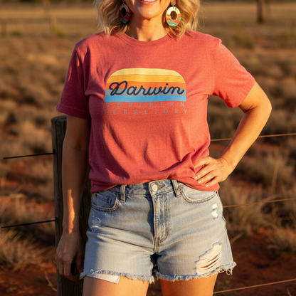 A woman wearing a coral pink graphic t-shirt with 'Darwin Australia' printed on the front, paired with light-wash denim shorts, posing outdoors in a rural setting with dry grass and a tree in the background.