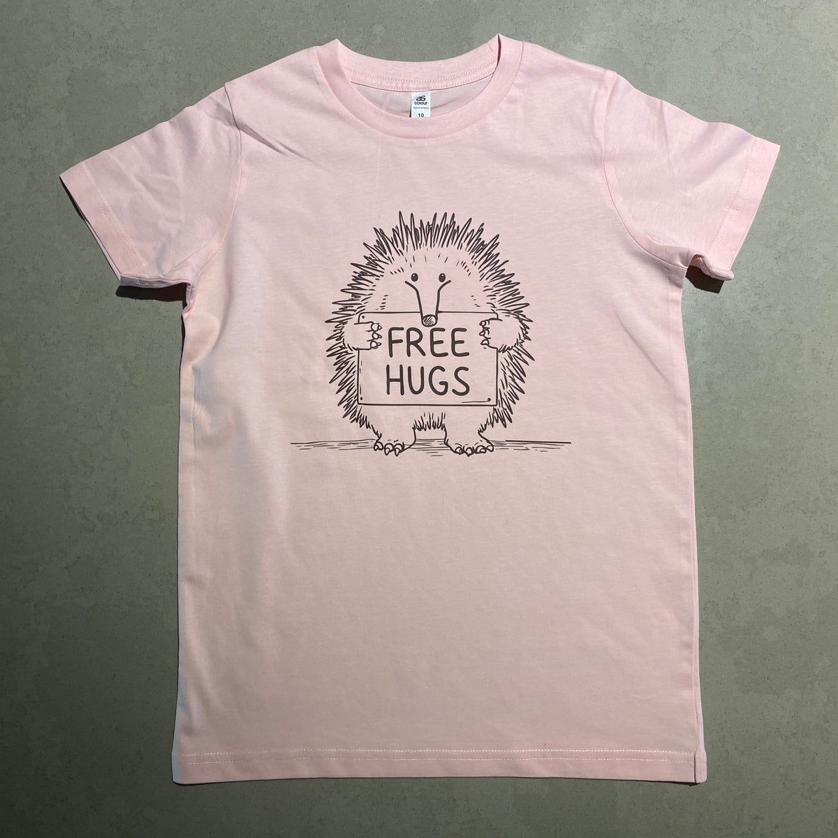 A light pink short-sleeve t-shirt laid flat on a gray-green textured surface. The front of the shirt features a black graphic of an echidna inside a circular frame with the text 'FREE HUGS'.
