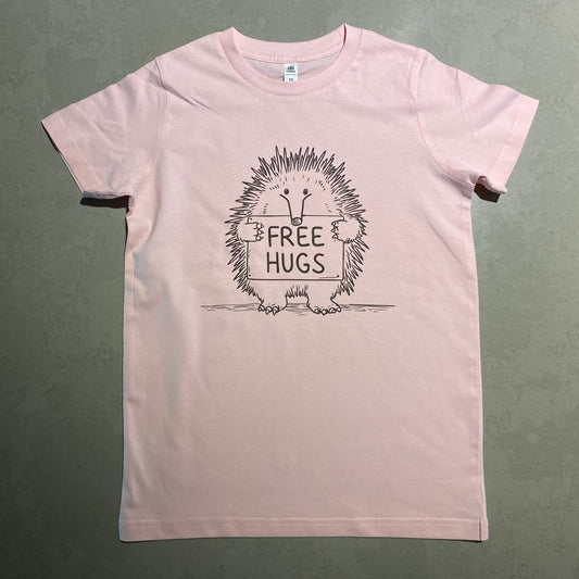 A light pink short-sleeve t-shirt laid flat on a gray-green textured surface. The front of the shirt features a black graphic of an echidna inside a circular frame with the text 'FREE HUGS'.
