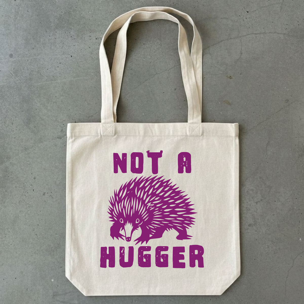A natural canvas tote bag with purple printed text reading 'NOT A HUGGER' and a purple hedgehog graphic, displayed flat on a gray concrete surface.