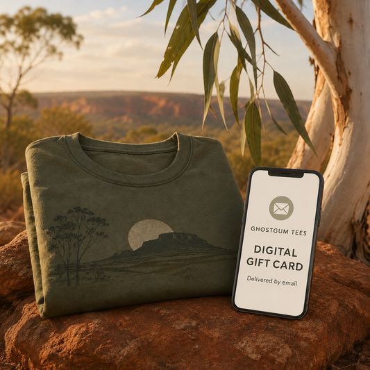 A folded olive green t-shirt with a landscape graphic is displayed outdoors on a rock next to a smartphone showing a Ghostgum Tees digital gift card, with a warm sunset background and tree branches.