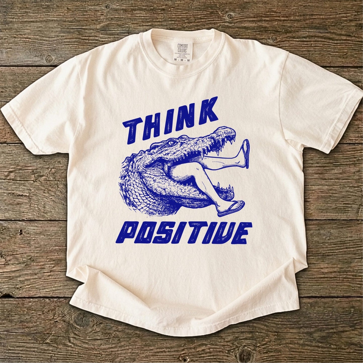 Folded t-shirt with blue saltwater croc graphic and 'THINK Positive' text on a white background