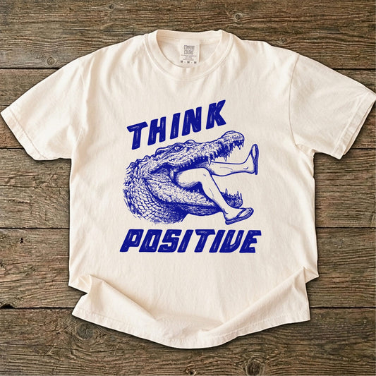 Folded t-shirt with blue saltwater croc graphic and 'THINK Positive' text on a white background