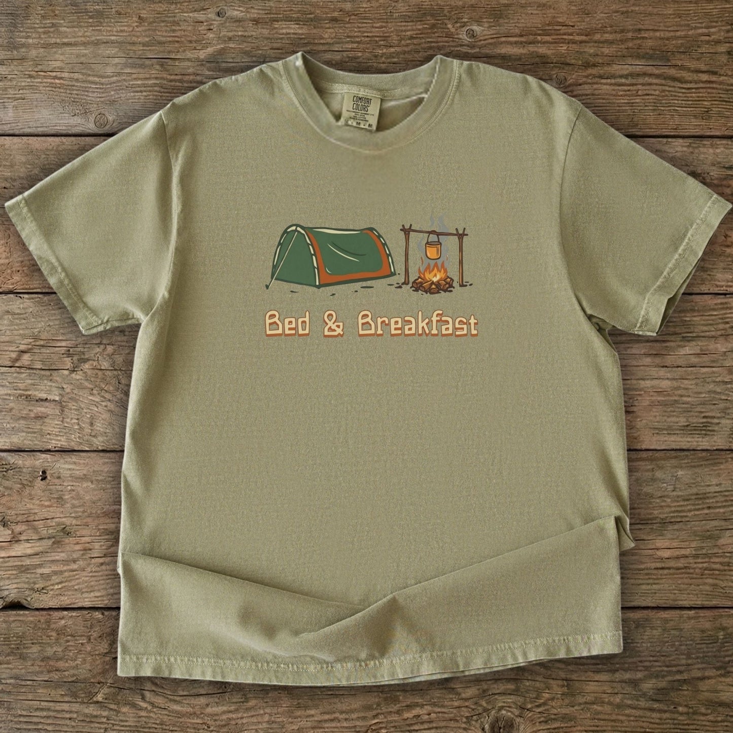 Green t-shirt with camping design and 'Bed & Breakfast' text on a wooden surface