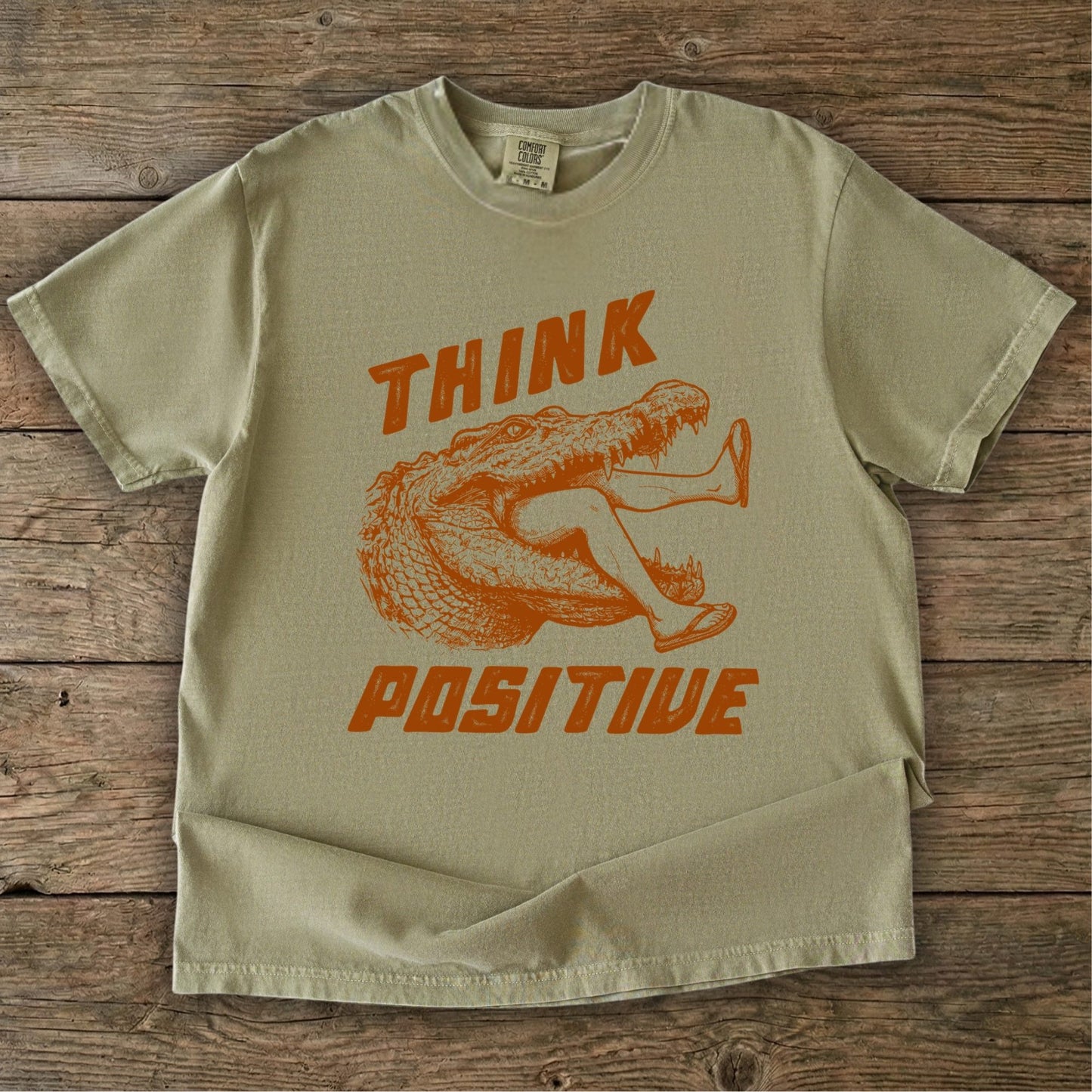 Folded t-shirt with saltwater croc graphic and 'THINK Positive' text on a wooden background