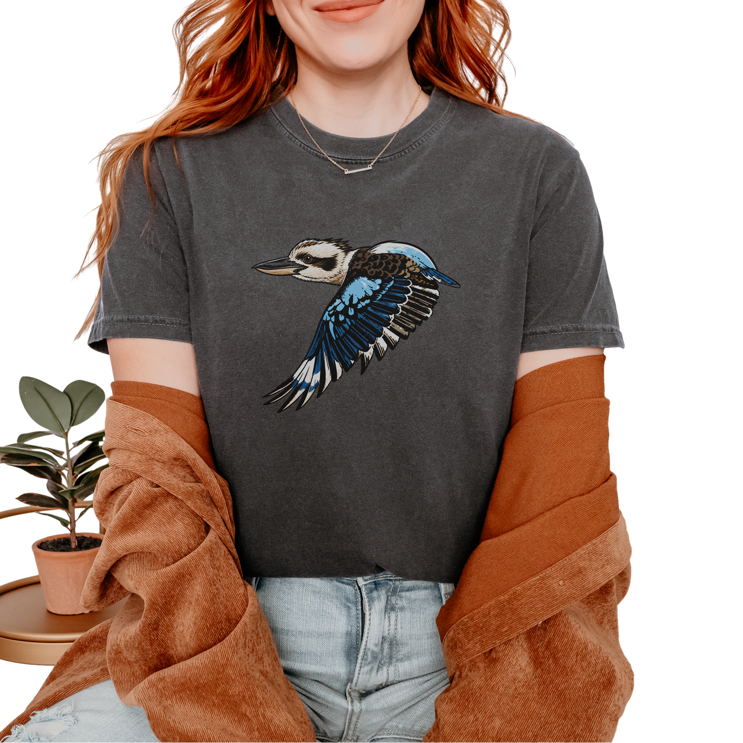 A woman wearing a dark gray graphic t-shirt with a flying kookaburra design, styled with a brown cardigan and light wash jeans against a white background.