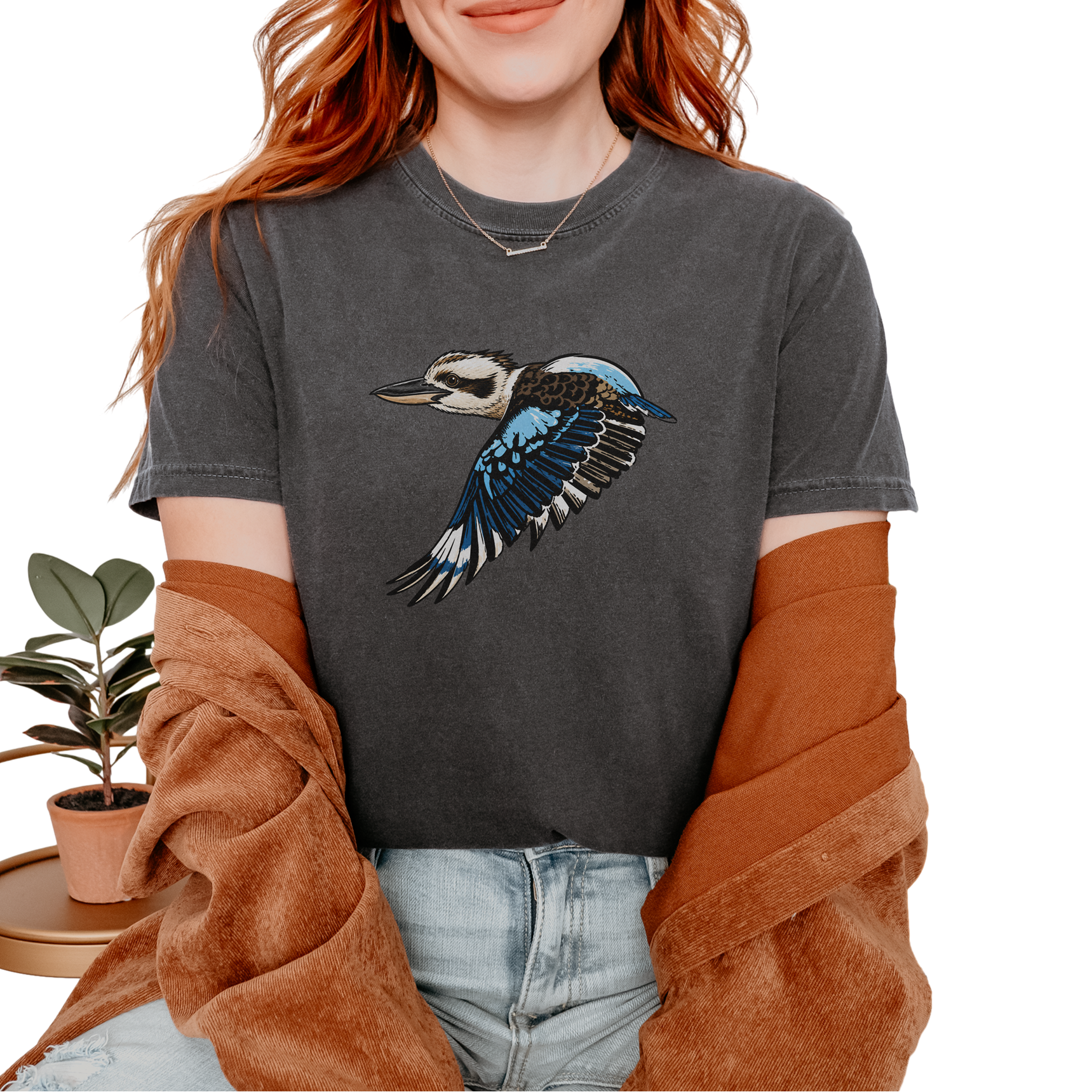 A woman wearing a dark gray graphic t-shirt with a flying kookaburra design, styled with a brown cardigan and light wash jeans against a white background.