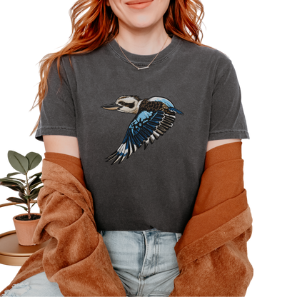 A woman wearing a dark gray graphic t-shirt with a flying kookaburra design, styled with a brown cardigan and light wash jeans against a white background.
