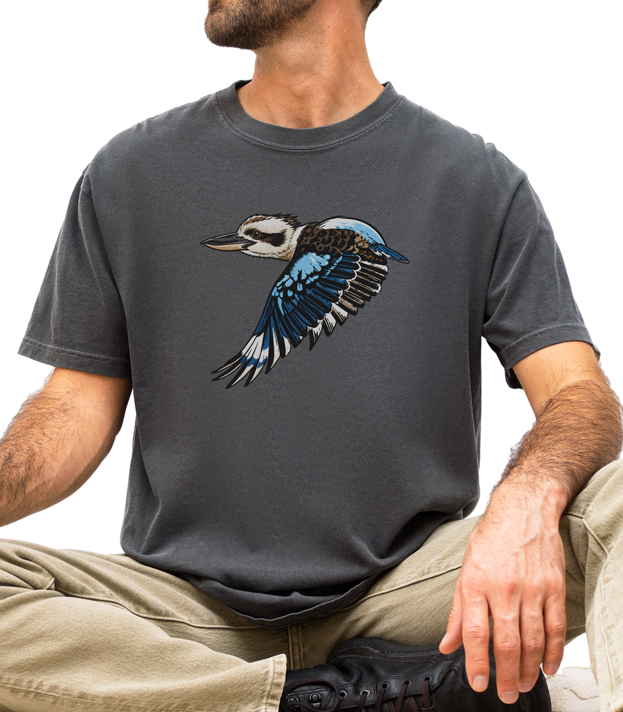 A man wearing a dark gray crewneck t-shirt with a graphic of a flying kookaburra bird, paired with khaki pants and black shoes, sitting cross-legged against a white background.