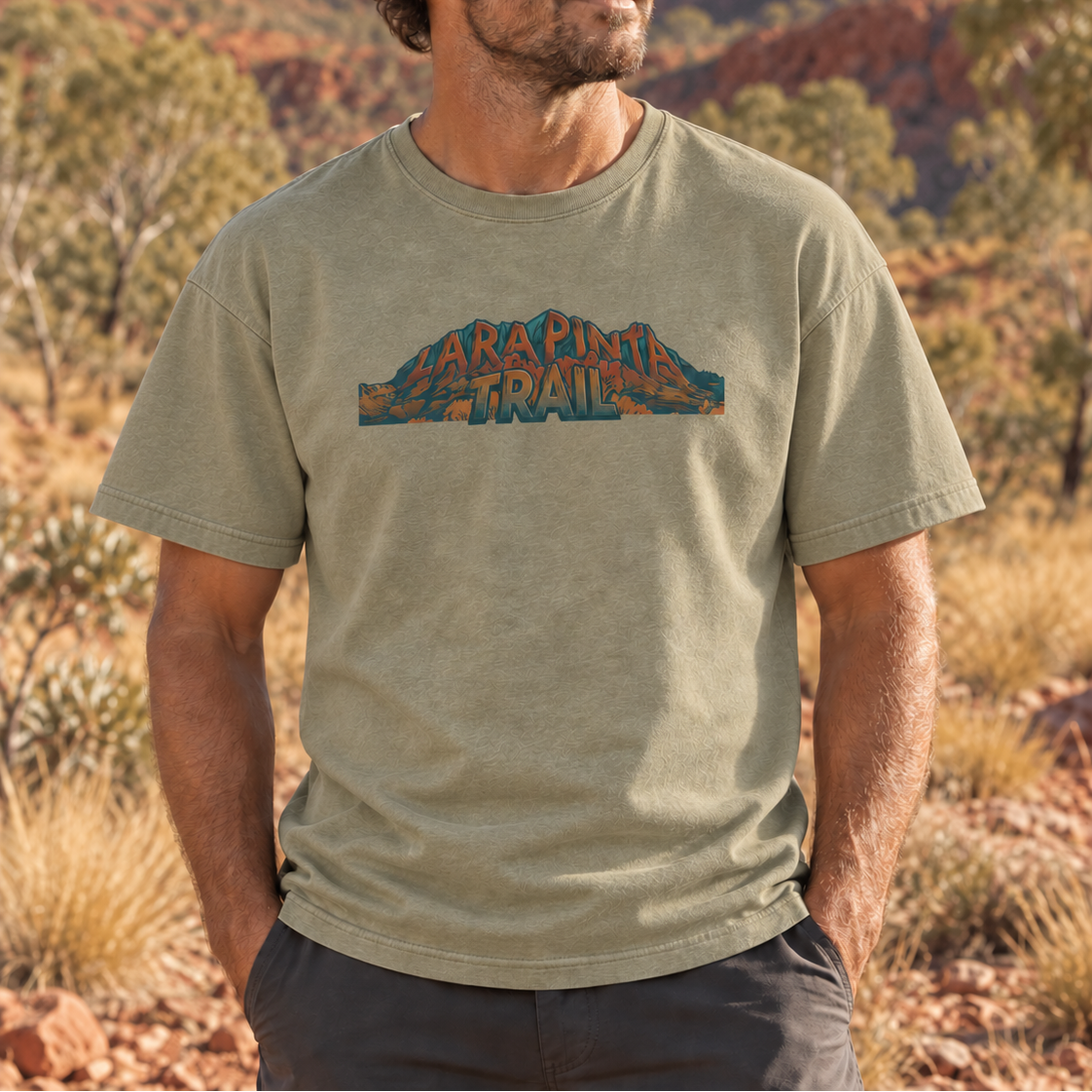 A man wearing a light olive green t-shirt with a 'Larapinta Trail' graphic on the chest, standing in a desert landscape with red rock mountains and trees in the background.