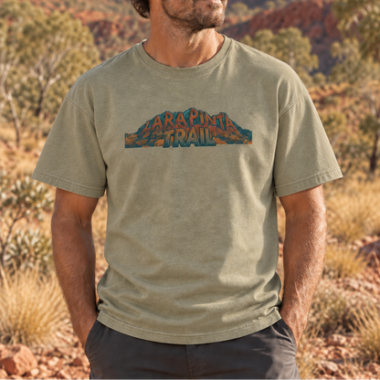 A man wearing a light olive green t-shirt with a 'Larapinta Trail' graphic on the chest, standing in a desert landscape with red rock mountains and trees in the background.