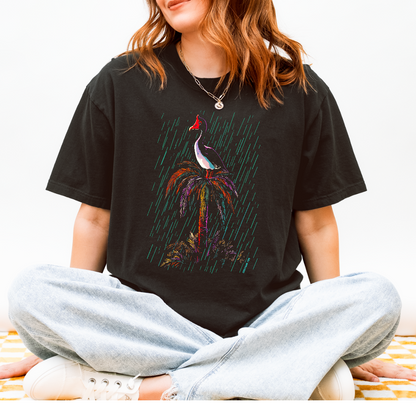 A woman sitting cross-legged wearing a black graphic t-shirt with a colorful Magpie Goose  bird print, light wash jeans, and white sneakers against a plain white background.