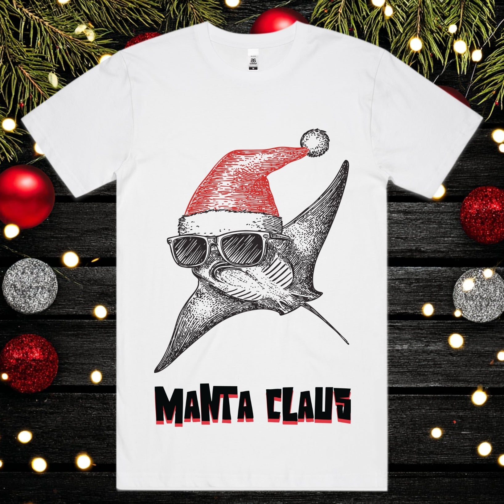 White t-shirt with a manta ray wearing a Santa hat and sunglasses, labeled 'Manta Claus', on a festive background with Christmas decorations.