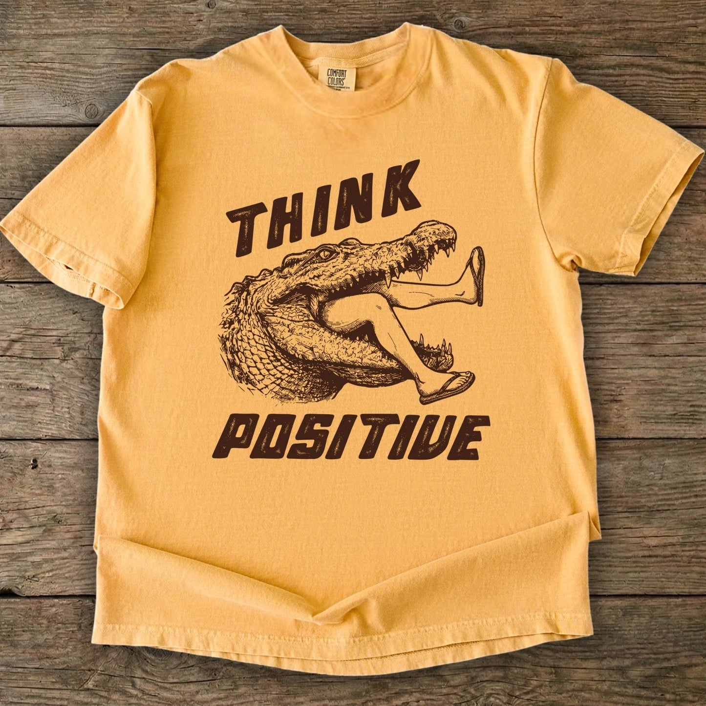 Folded t-shirt with saltwater croc graphic and 'THINK Positive' text on a wooden background