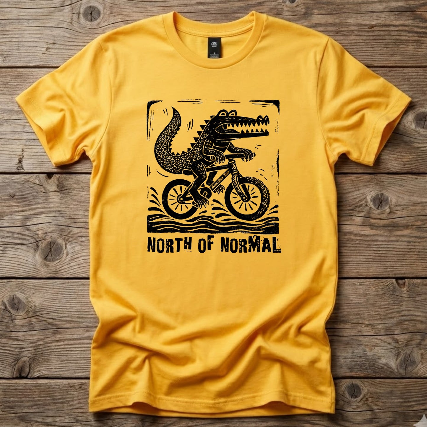 Yellow Ghost Gum Tees t-shirt featuring a black linocut-style graphic of a crocodile riding a bicycle with the text North of Normal.