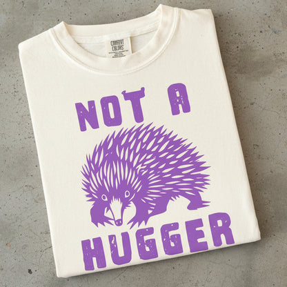 An off-white Comfort Colors t-shirt folded flat, featuring a purple graphic of an echidna with the text 'NOT A HUGGER' printed in purple.