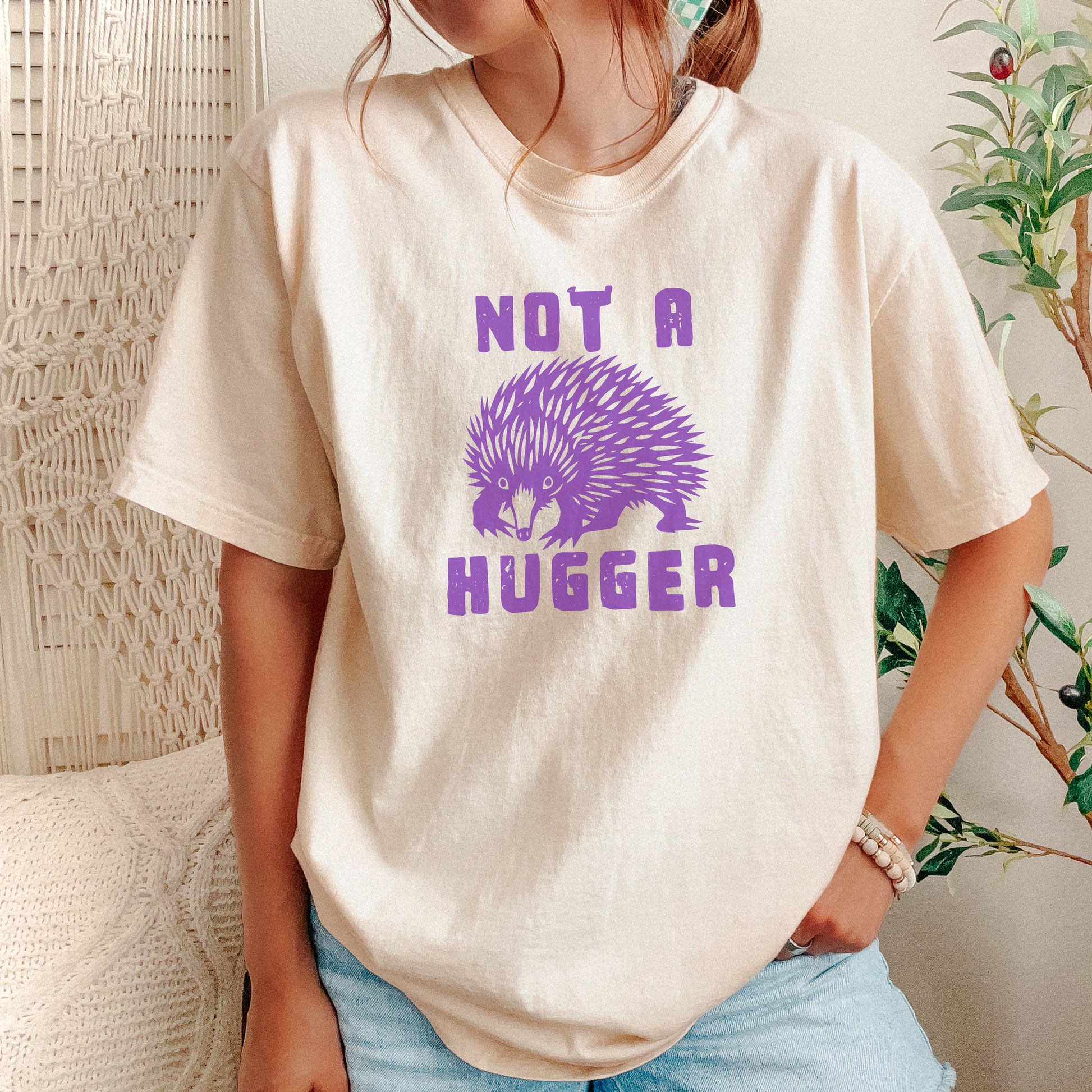 A person wearing a cream-colored t-shirt with a purple graphic of an echidna and the text 'NOT A HUGGER'. The person is also wearing light blue denim shorts, standing in a cozy indoor setting with a macrame wall hanging and a potted plant in the background.