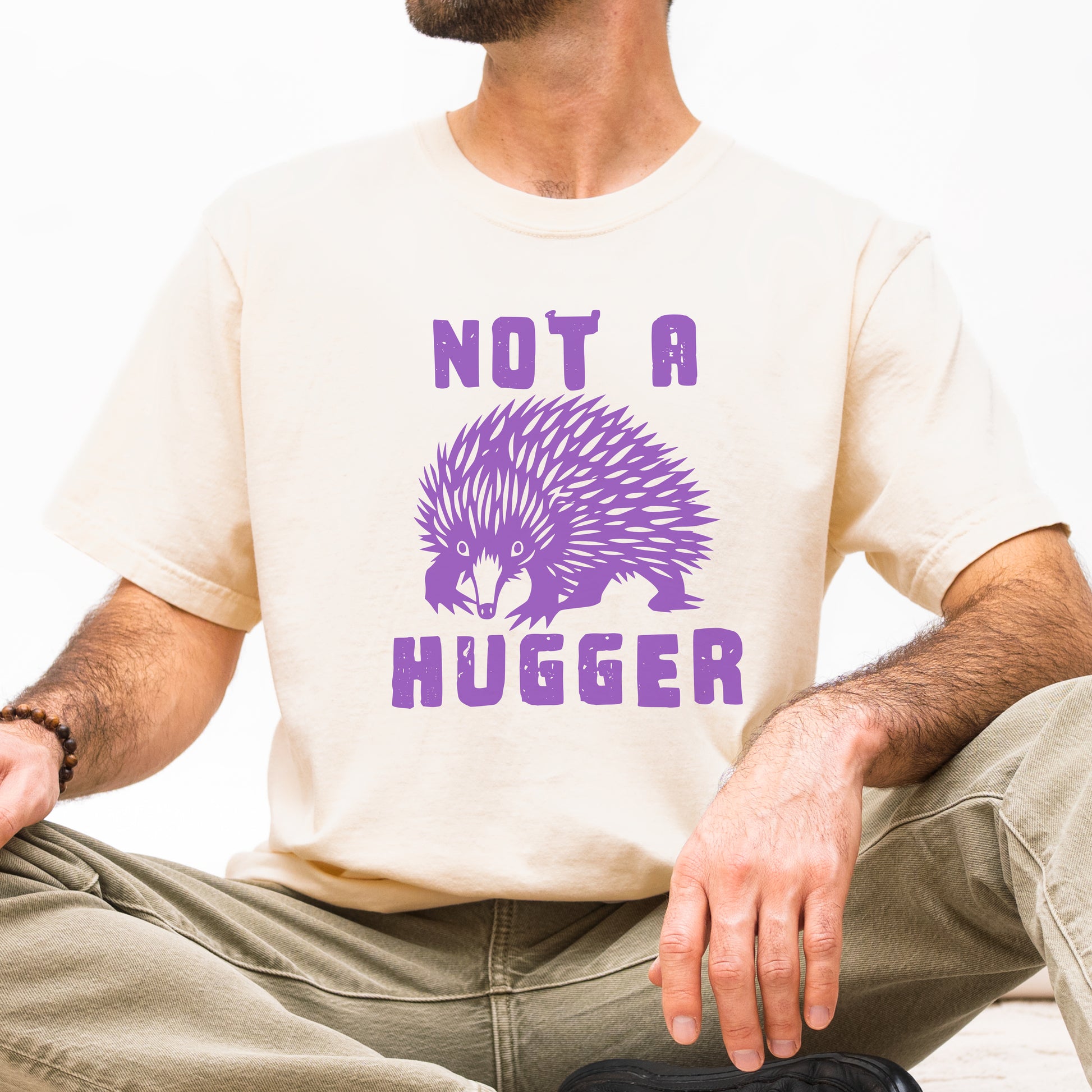 A man wearing a cream-colored t-shirt with a purple graphic of an echidna and the text 'NOT A HUGGER' in purple, paired with khaki pants.