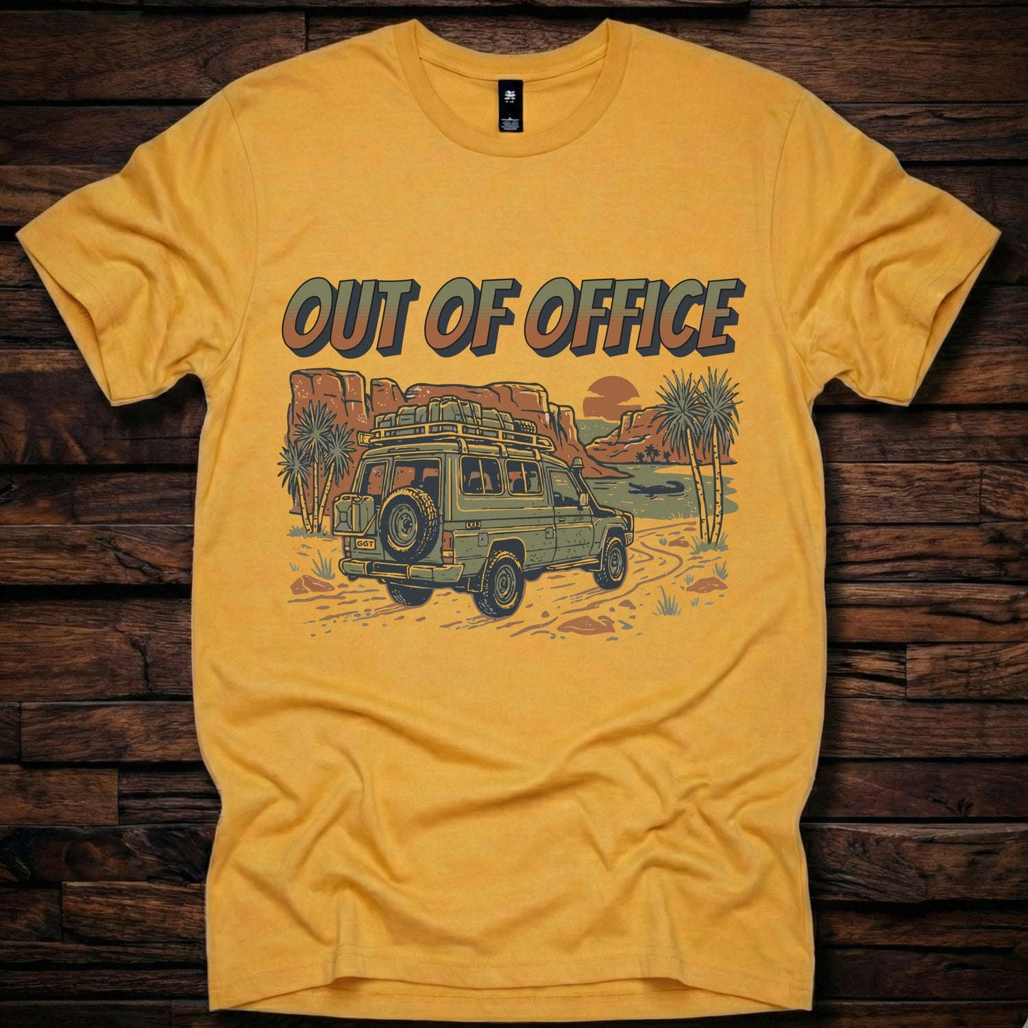 Mustard yellow t-shirt featuring a retro 'Out of Office' graphic with a green 4x4 off-road vehicle and Australian outback camping scene on a rustic wooden background.