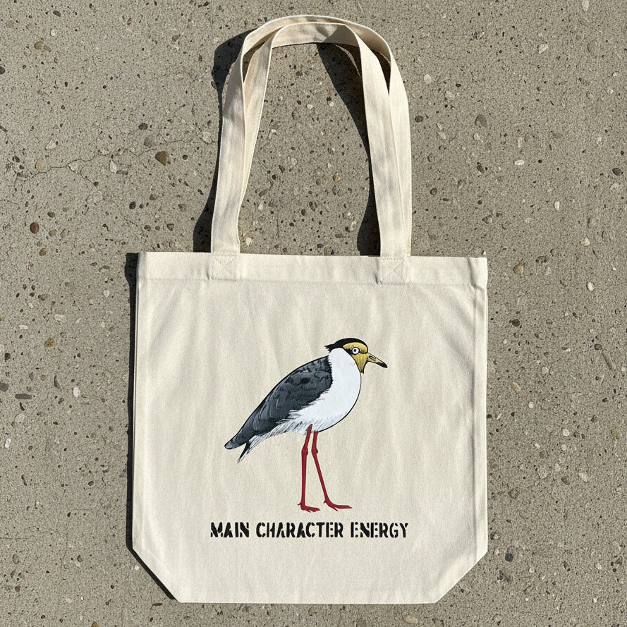 A natural canvas tote bag with a printed illustration of a plover bird and the text 'MAIN CHARACTER ENERGY' laid flat on a light wood surface.