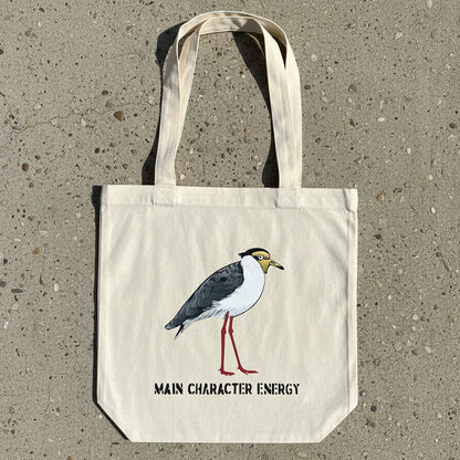 A natural canvas tote bag with a printed illustration of a plover bird and the text 'MAIN CHARACTER ENERGY' laid flat on a light wood surface.