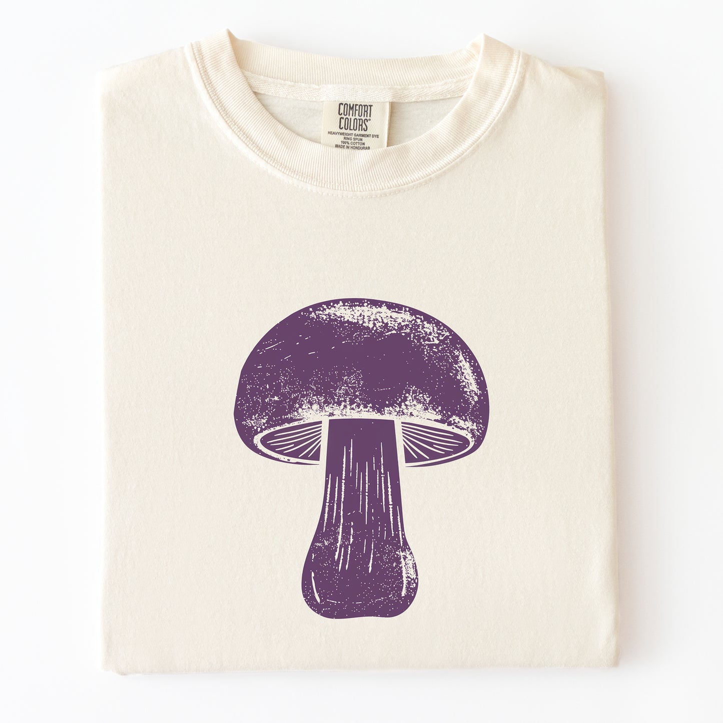 A folded cream-colored t-shirt with a distressed purple mushroom graphic printed on the front, displayed against a clean white background.