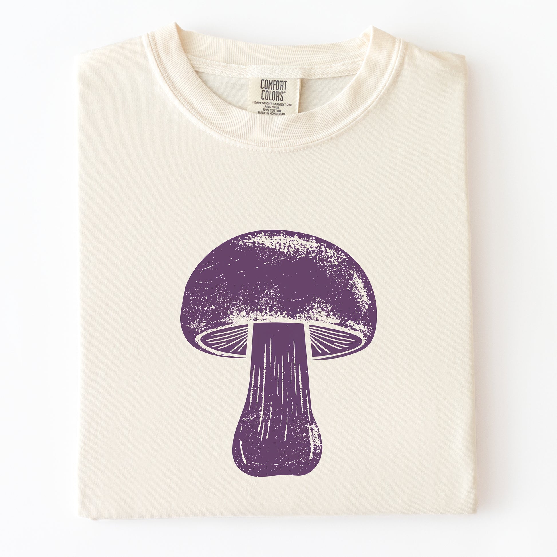A folded cream-colored t-shirt with a distressed purple mushroom graphic printed on the front, displayed against a clean white background.