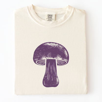 A folded cream-colored t-shirt with a distressed purple mushroom graphic printed on the front, displayed against a clean white background.
