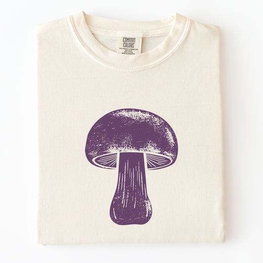 A folded cream-colored t-shirt with a distressed purple mushroom graphic printed on the front, displayed against a clean white background.