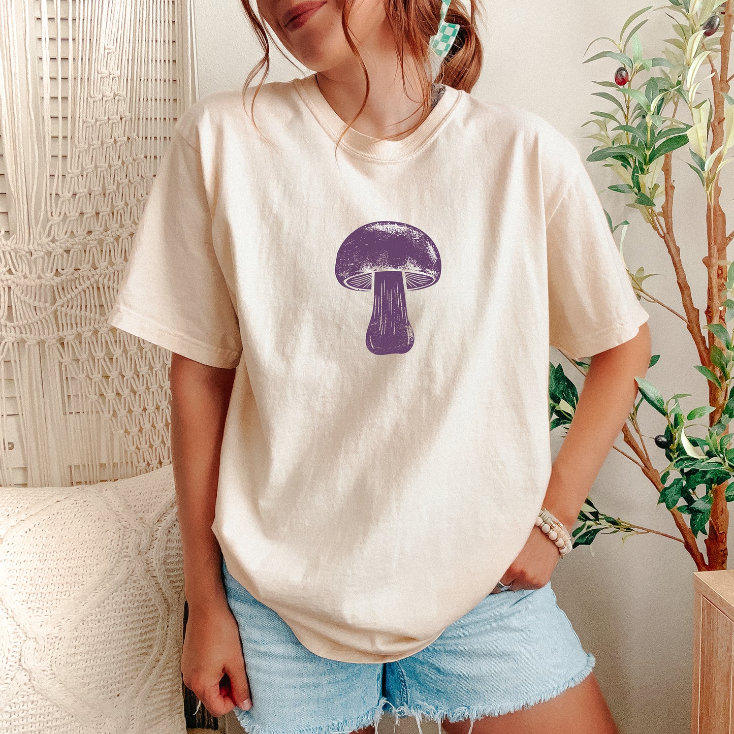 A woman wearing a cream-colored t-shirt with a purple mushroom graphic on the chest, paired with light blue denim shorts, standing in a cozy room setting with a macrame wall hanging and a potted plant.