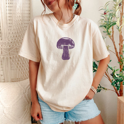 A woman wearing a cream-colored t-shirt with a purple mushroom graphic on the chest, paired with light blue denim shorts, standing in a cozy room setting with a macrame wall hanging and a potted plant.