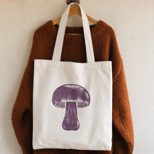 A white canvas tote bag with a purple mushroom graphic, hanging on a wooden hanger from a brown knit sweater against a textured off-white wall.