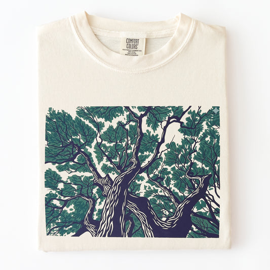 A folded cream-colored Comfort Colors t-shirt featuring a large graphic print of a gnarled tree with dark green and navy blue leaves on the front, set against a plain white background.