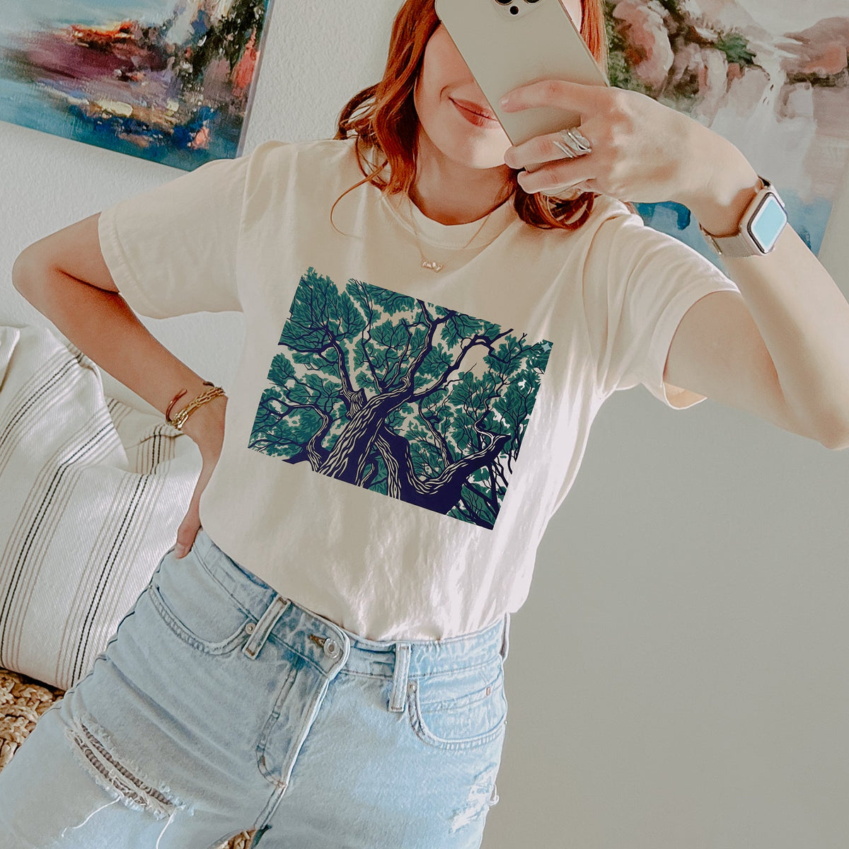 A woman takes a mirror selfie wearing a cream-colored t-shirt with a large green and blue tree graphic on the front, paired with light-wash distressed jeans.